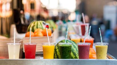 Six gobelets en plastique transparent remplis de jus de fruits colorés se trouvent devant divers fruits.